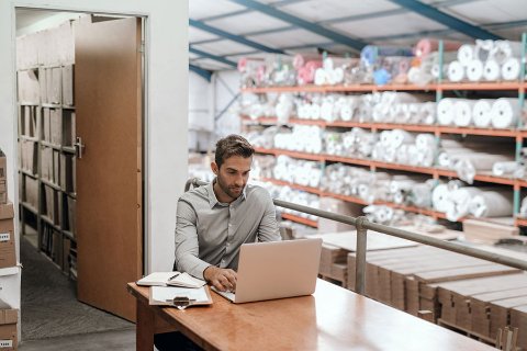 Homme d'affaires travaillant sur un ordinateur portable, avec des rouleaux de tissu en arrière-plan, représentant un stockage pour entreprise du secteur de la mode